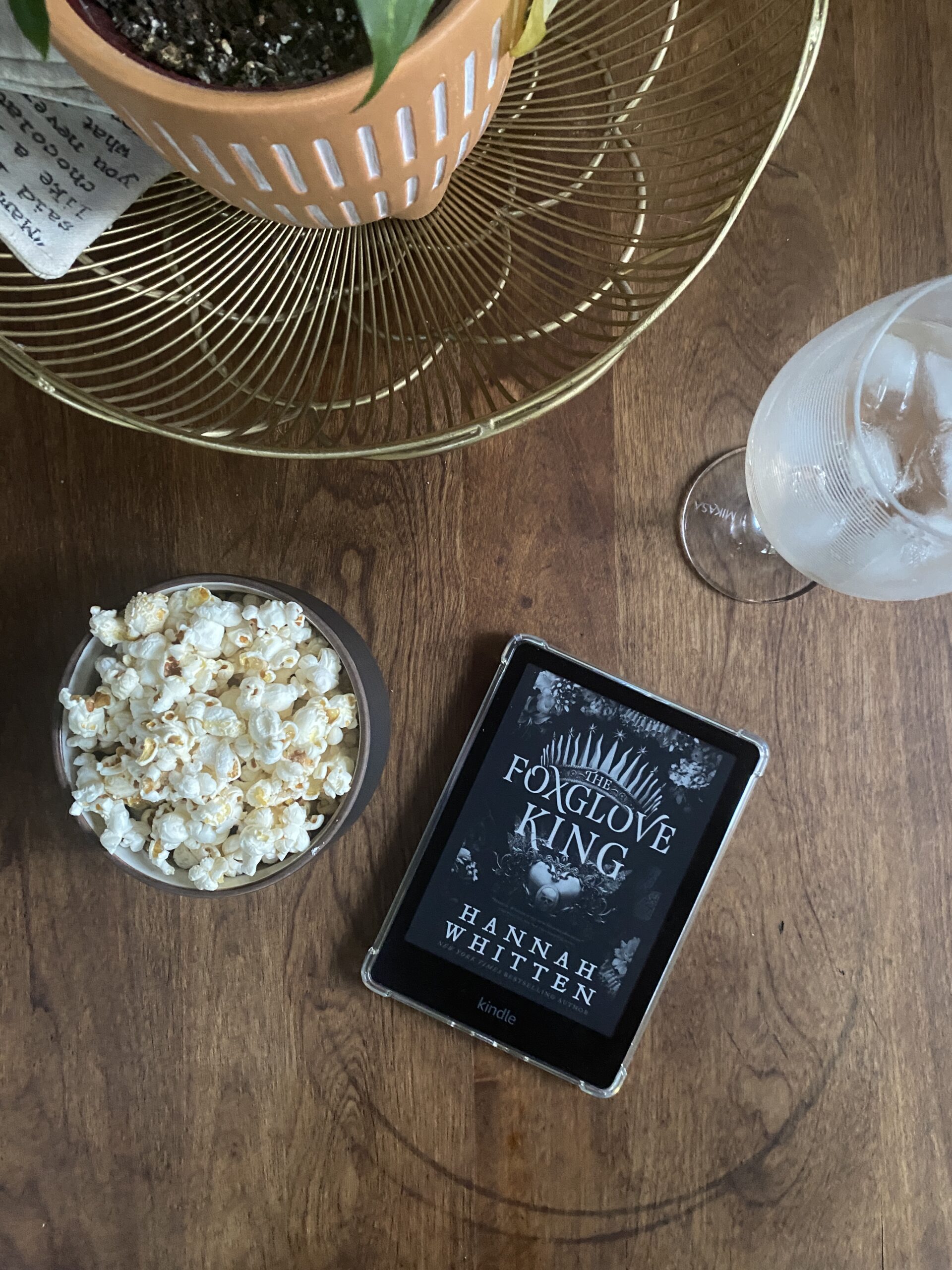 Kindle reader with The Foxglove King book cover beside a bowl of popcorn and glass of white wine